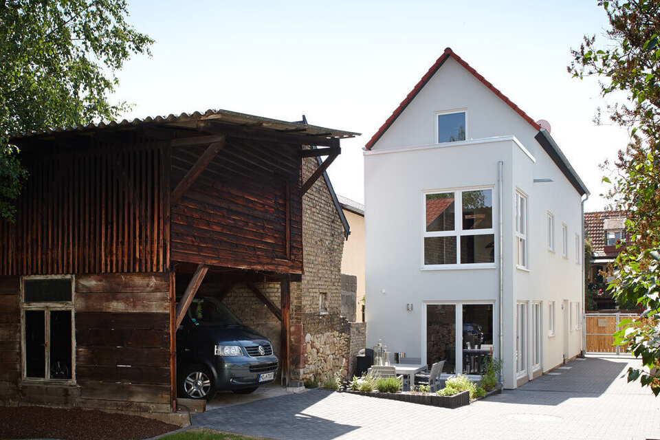 Ein modernes, zweigeschossiges Haus mit großen Fenstern neben einem traditionellen Holzgebäude. Ein Auto steht in der Einfahrt, umgeben von grünem Rasen und Bäumen.