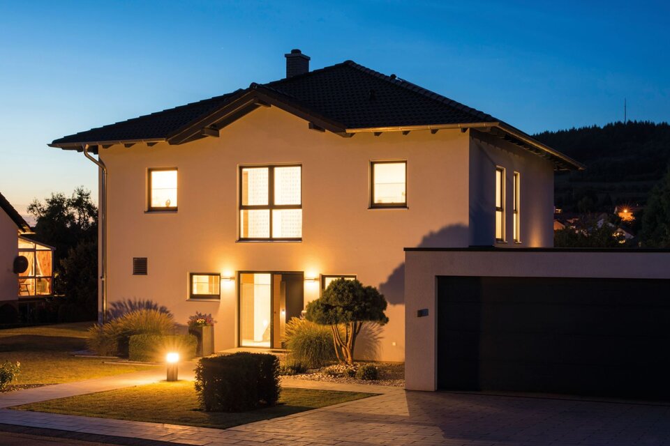 Modernes zweigeschossiges FingerHaus bei Dämmerung mit beleuchtetem Eingangsbereich, dunklem Satteldach, großer Garage und gepflegtem Vorgarten.