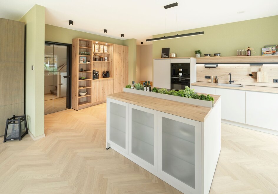 Große, moderne Küche mit zentraler Kochinsel, weißen Fronten und Holzarbeitsplatte im Musterhaus JUNO Mülheim-Kärlich. Grüne Wandfarbe, offene Regale und Glasvitrine verleihen dem Raum eine warme, natürliche Atmosphäre.