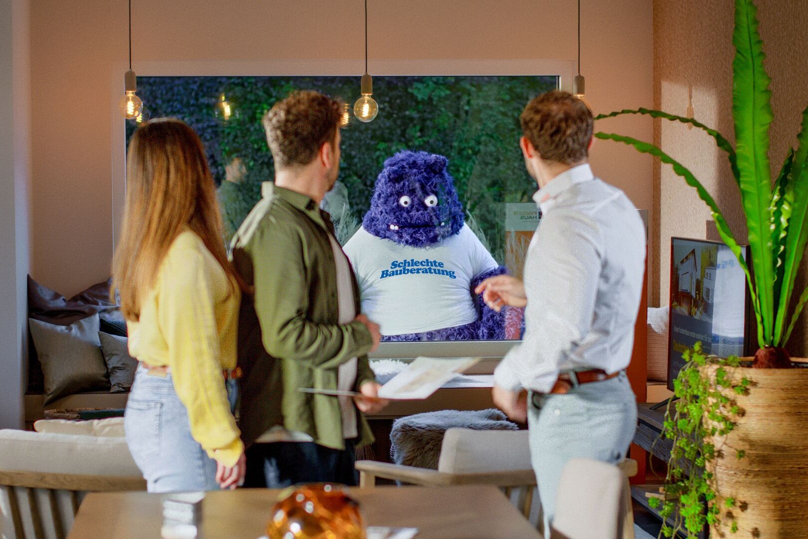 Drei Personen stehen in einem modern eingerichteten Wohnzimmer und blicken durch das große Fenster auf ein blaues, plüschartiges Wesen mit der Aufschrift „Schlechte Bauberatung“ – Symbolfigur aus der FingerHaus-Kampagne „Das Unerwartete“.