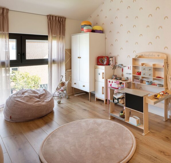 Kinderzimmer mit Spielküche, Spielzeug, Teppich und Sitzsack neben einem großen Fenster