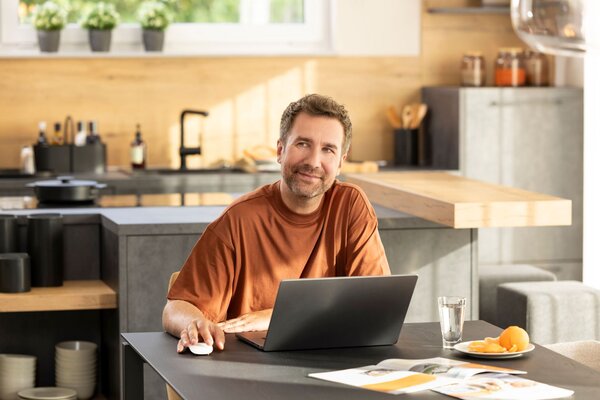 Interessent sitzt an einem Tisch mit Laptop in einer offenen Küche, blickt nachdenklich zur Seite; auf dem Tisch liegen Unterlagen, ein Glas Wasser und ein Teller mit Obst.
