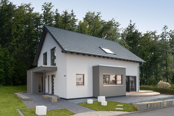 Modernes Musterhaus mit grauem Satteldach und zweifarbiger Putzfassade in Weiß und Grau. Der Eingangsbereich ist überdacht, rechts schließt eine Holzterrasse an. Im Vordergrund sind gepflasterte Wege und Rasenflächen zu sehen.