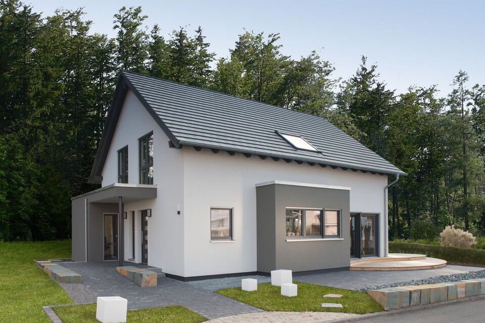 Modernes Musterhaus mit grauem Satteldach und zweifarbiger Putzfassade in Weiß und Grau. Der Eingangsbereich ist überdacht, rechts schließt eine Holzterrasse an. Im Vordergrund sind gepflasterte Wege und Rasenflächen zu sehen.