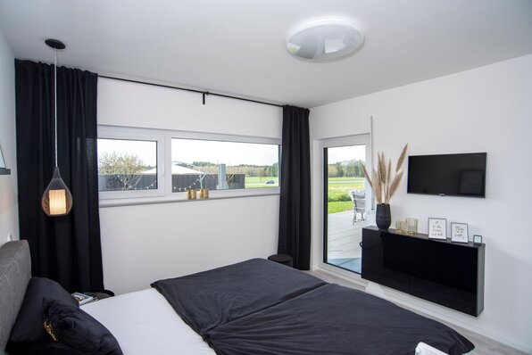 Schlafzimmer in einem FingerHaus NIVO 130 mit großem Bett, schwarzer Bettwäsche, breiter Fensterfront, Zugang zur Terrasse und moderner Dekoration.