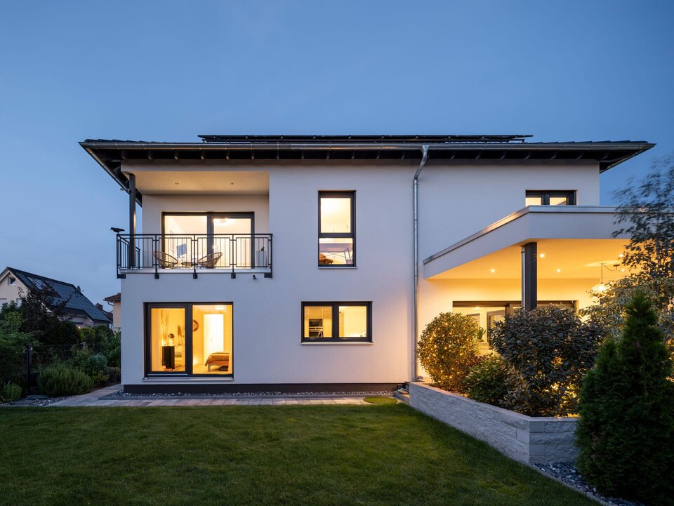 Rückansicht der frei geplanten FingerHaus Stadtvilla in der Abenddämmerung mit Balkon, überdachter Terrasse und beleuchteten Innenräumen.