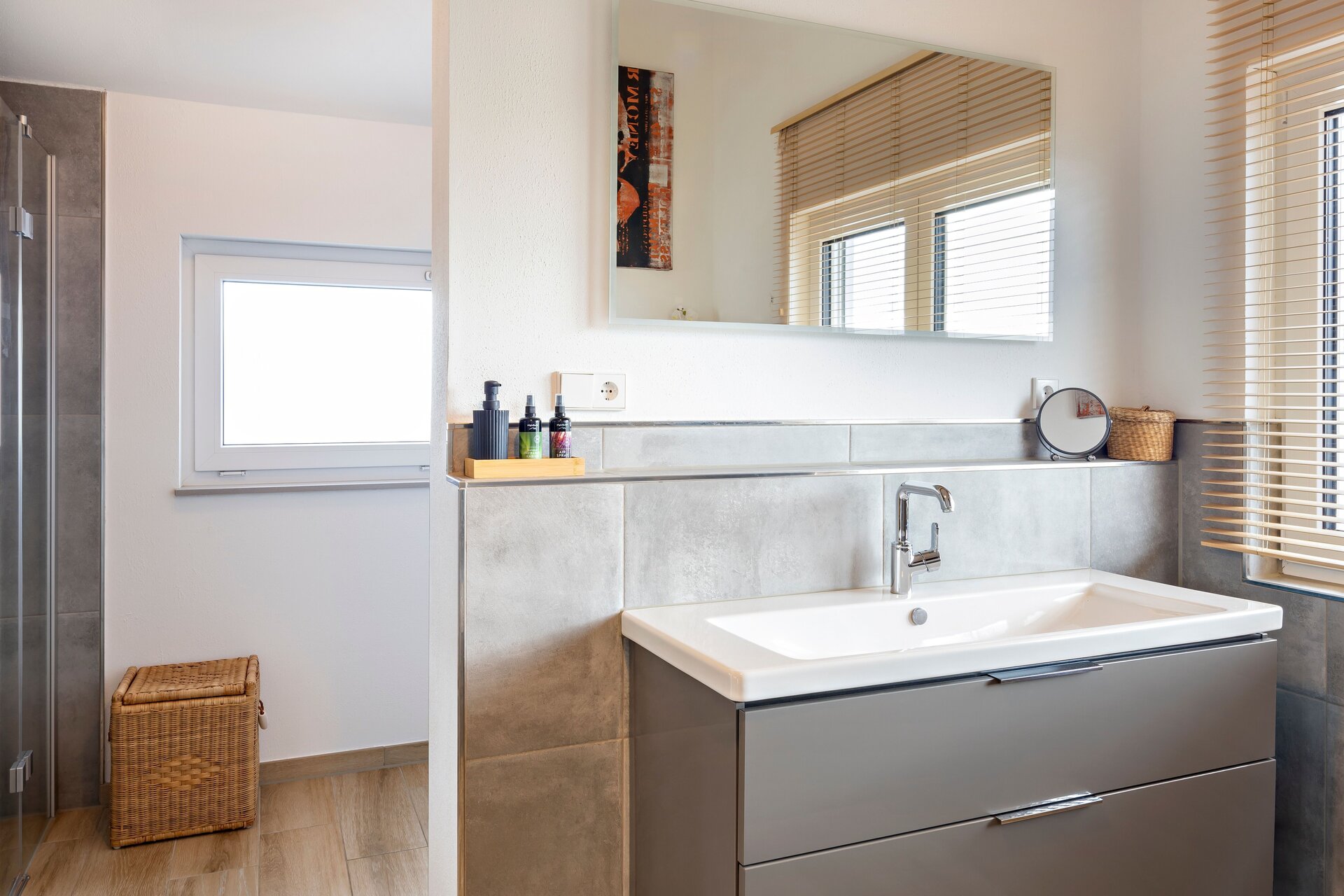 Badezimmer mit grauem Waschtischunterschrank, weißem Waschbecken, Wandspiegel, grauen Wandfliesen und bodengleicher Dusche.