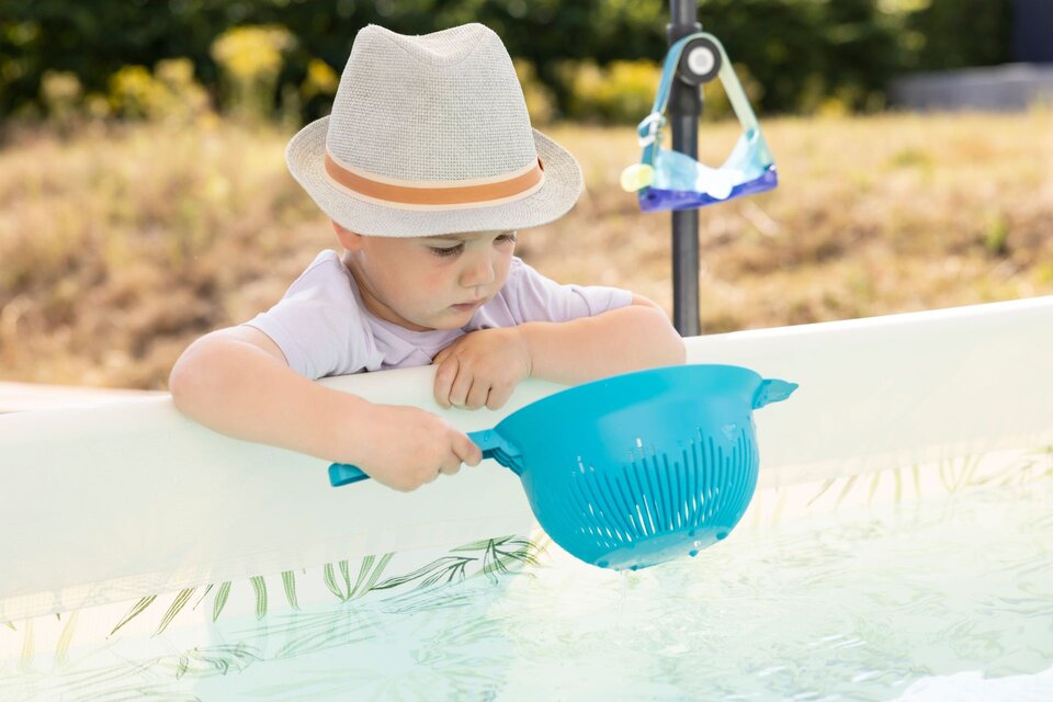 Kind mit Hut hält ein blaues Sieb über einem Wasserbecken im Garten und blickt ins Wasser