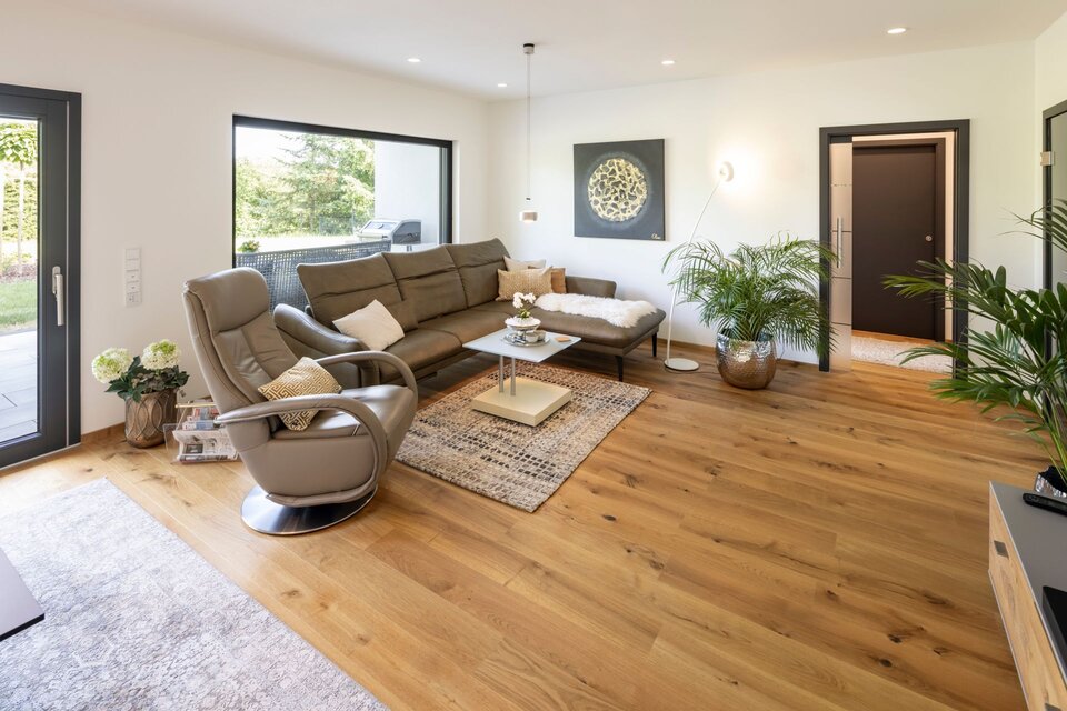 Wohnzimmer eines frei geplanten FingerHaus mit großem Ecksofa, modernem Relaxsessel und Holzboden. Helle Wände, große Fenster und stilvolle Pflanzen sorgen für ein freundliches Ambiente.