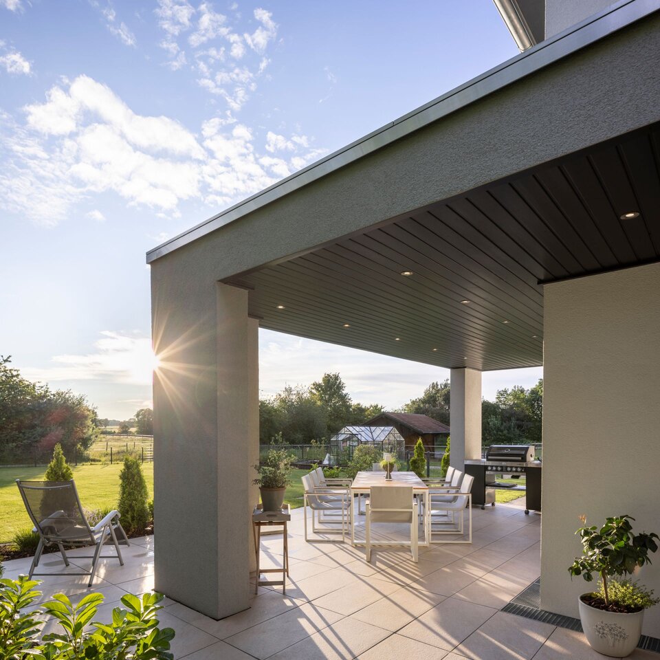 Moderne, überdachte Terrasse eines FingerHaus-Hauses bei tiefstehender Sonne mit stilvollen Gartenmöbeln, gepflegtem Rasen und Blick auf ein Gewächshaus im Hintergrund.