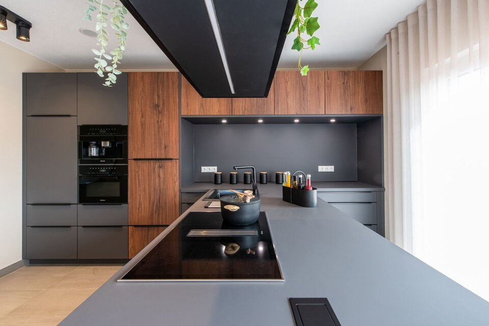 Moderne Küche mit großer Kochinsel, grauer Arbeitsplatte und Fronten in Holzoptik im Musterhaus TALO Babenhausen. Schwarze Einbaugeräte, Dunstabzug im Deckenelement und hängende Pflanzen verleihen der Küche ein stilvolles Ambiente.