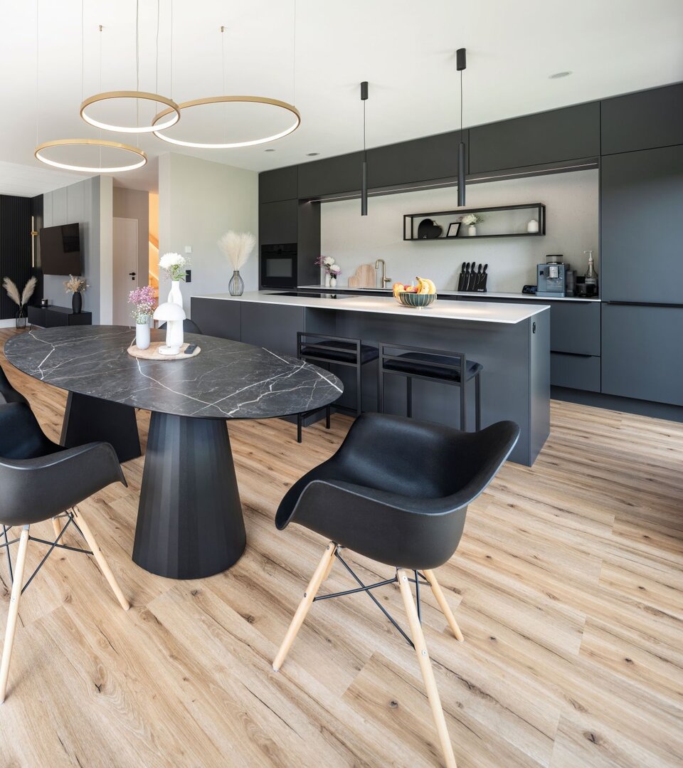 Moderne offene Wohnküche mit schwarzer Einbauküche, rundem Esstisch aus dunklem Stein, vier schwarzen Stühlen und hellen Holzboden. Im Hintergrund Wohnbereich mit Sofa und Couchtisch, große Fensterfront und drei runde Pendelleuchten über dem Tisch.
