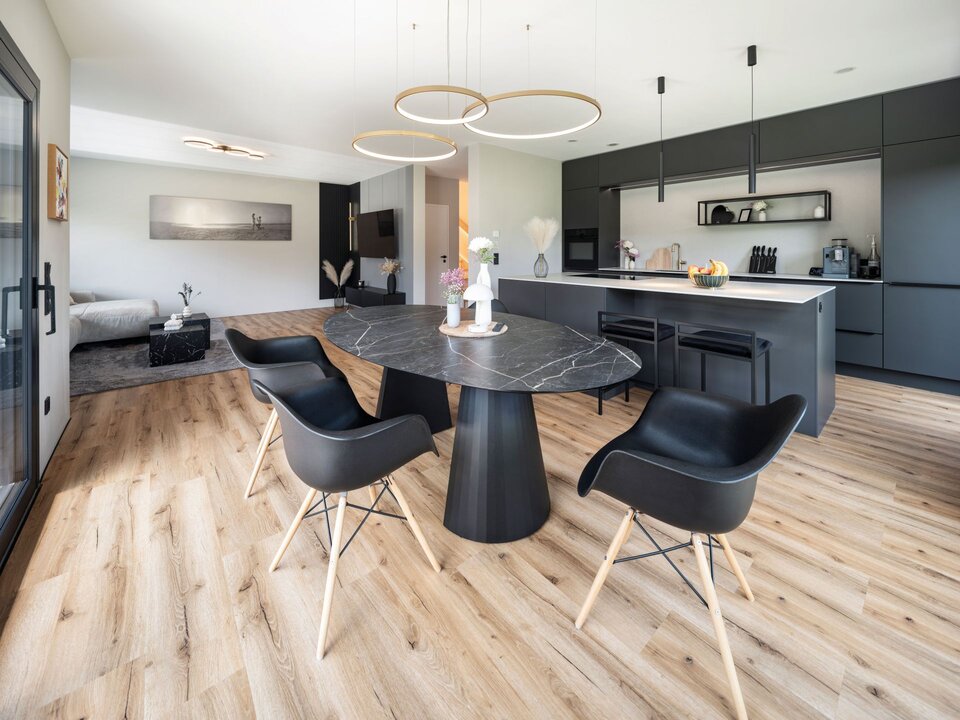 Moderne offene Wohnküche mit schwarzer Einbauküche, rundem Esstisch aus dunklem Stein, vier schwarzen Stühlen und hellen Holzboden. Im Hintergrund Wohnbereich mit Sofa und Couchtisch, große Fensterfront und drei runde Pendelleuchten über dem Tisch.