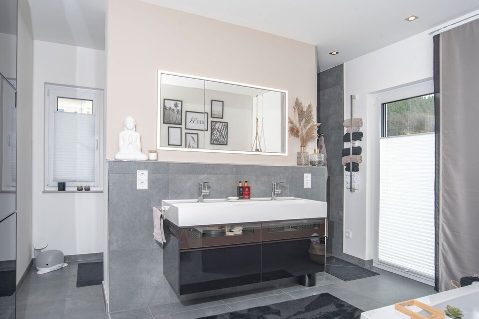 Modernes Badezimmer in einem FingerHaus NIVO 130 mit großem Doppelwaschtisch, beleuchtetem Spiegel, grauen Fliesen, dekorativen Accessoires und zwei Fenstern mit Sichtschutz.