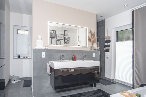 Modernes Badezimmer in einem FingerHaus NIVO 130 mit großem Doppelwaschtisch, beleuchtetem Spiegel, grauen Fliesen, dekorativen Accessoires und zwei Fenstern mit Sichtschutz.