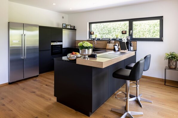 Moderne Küche in einem frei geplanten FingerHaus mit schwarzer Kochinsel, Holzarbeitsplatte und integriertem Tresen mit Barhockern. Große Fenster bringen viel Tageslicht in den Raum, ergänzt durch Edelstahl-Kühlschrank und hochwertige Elektrogeräte.