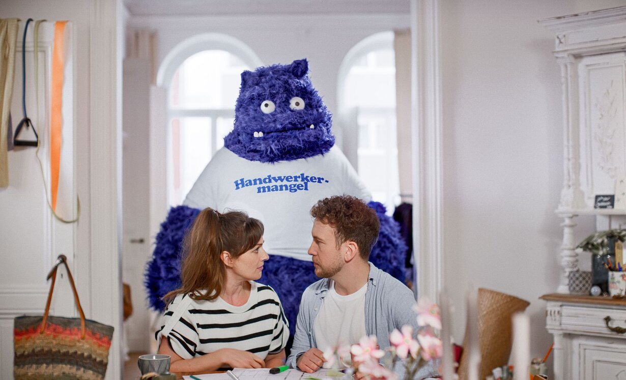 Ein Paar sitzt am Tisch und bespricht Baupläne, während hinter ihnen ein großes blaues, plüschartiges Wesen mit der Aufschrift „Handwerkermangel“ auf dem T-Shirt steht – Symbolfigur aus der Kampagne „Das Unerwartete“.