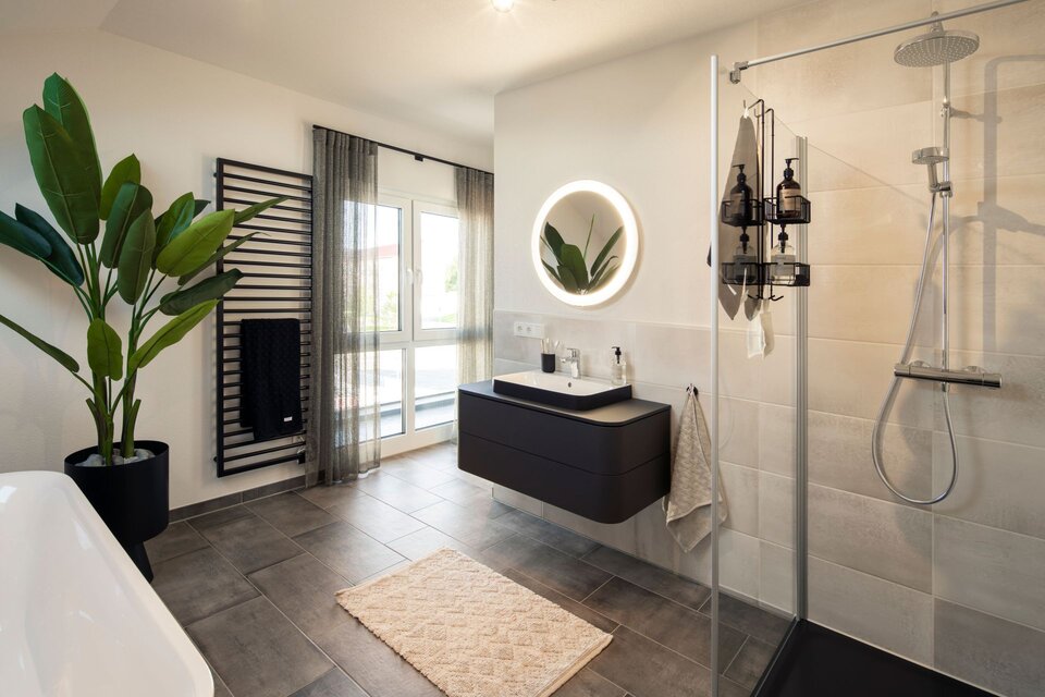 Modernes Badezimmer mit bodengleicher Dusche, freistehender Badewanne und großem Spiegel mit Beleuchtung. Dunkler Waschtisch, große Fliesen und Pflanzen sorgen für ein stilvolles Ambiente.