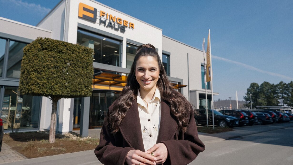 Sally Özcan, bekannte Influencerin, mit langen dunklen Haaren und dunklem Mantel steht vor dem Firmengebäude von FingerHaus mit sichtbarem Firmenlogo und mehreren geparkten Autos im Hintergrund.
