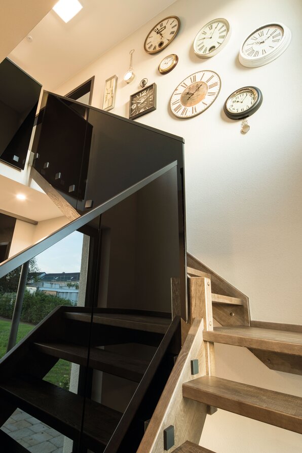 Moderne Holztreppe im Musterhaus MAXIM in Gießen mit dekorativer Uhrenwand und stilvollem schwarzen Geländer.