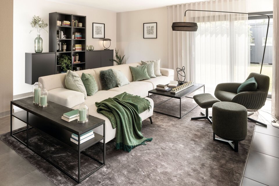 Modernes Wohnzimmer im FingerHaus NEO 300 Musterhaus Hannover mit großem hellen Sofa, grünen Akzenten, eleganter Einrichtung und viel Tageslicht durch große Fensterfront.