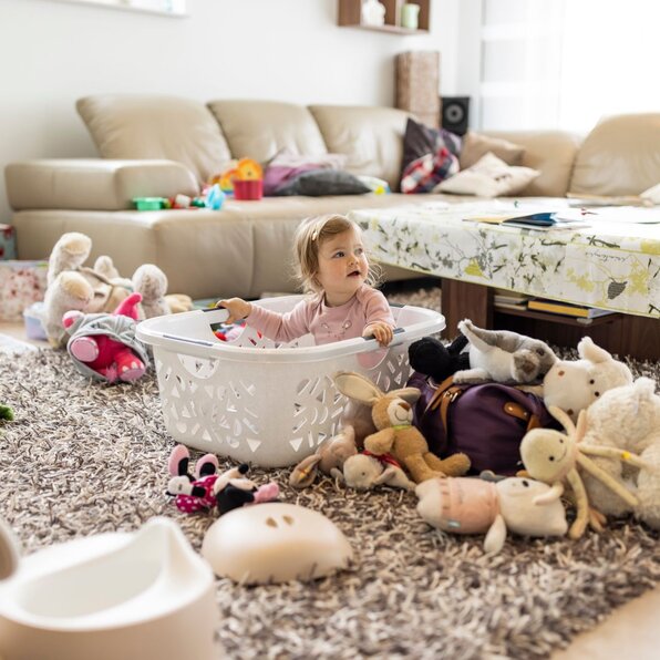 Ein kleines Kind sitzt in einem weißen Wäschekorb auf einem Teppich im Wohnzimmer, umgeben von zahlreichen Kuscheltieren und Spielsachen. Im Hintergrund steht ein beiger Ledersessel mit Tisch und Spielzeug darauf.