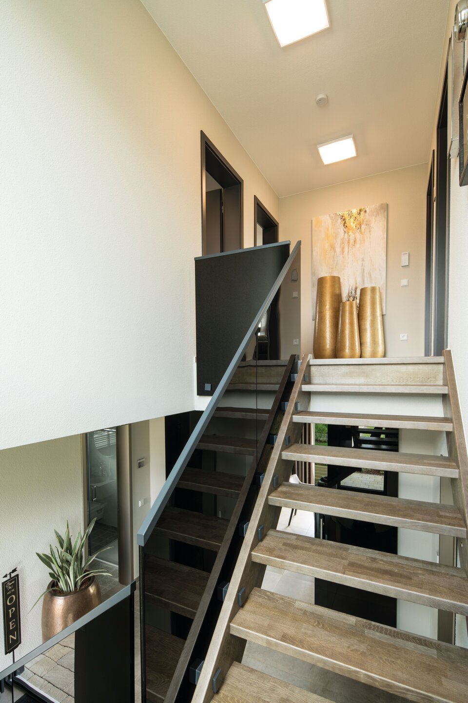 Moderner Innenraum mit offener Treppe aus Holzstufen und schwarzem Geländer, im Hintergrund zwei große goldfarbene Vasen auf einem weißen Sideboard unter einem Oberlicht