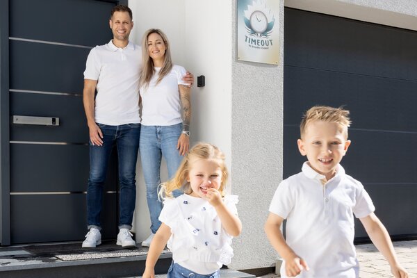 Zwei Kinder rennen lachend auf dem gepflasterten Weg vor einem modernen FingerHaus-Haus, während ihre Eltern an der Haustür stehen und ihnen lächelnd zusehen.