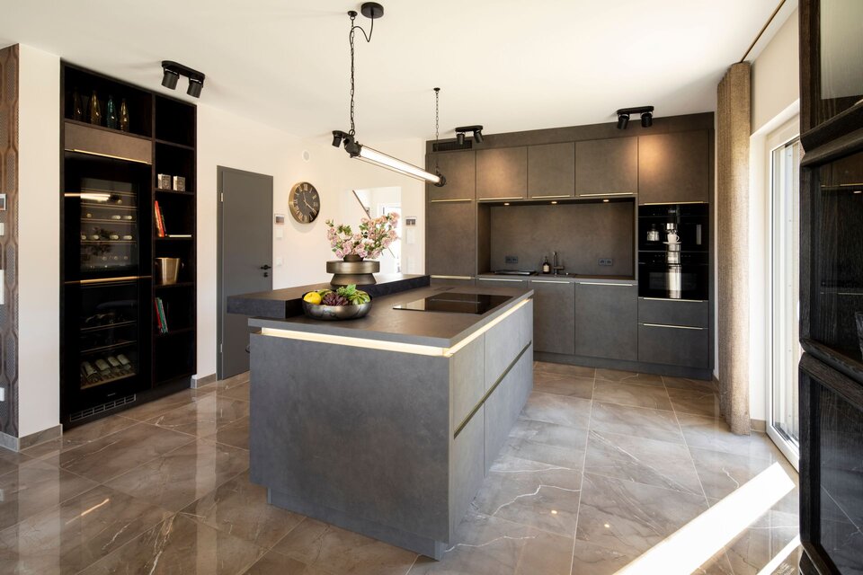 Moderne Küche in dunkler Betonoptik mit großer Kochinsel und integrierter Beleuchtung. Glasvitrine, schwarze Einbaugeräte und glänzender Fliesenboden betonen das elegante Design.