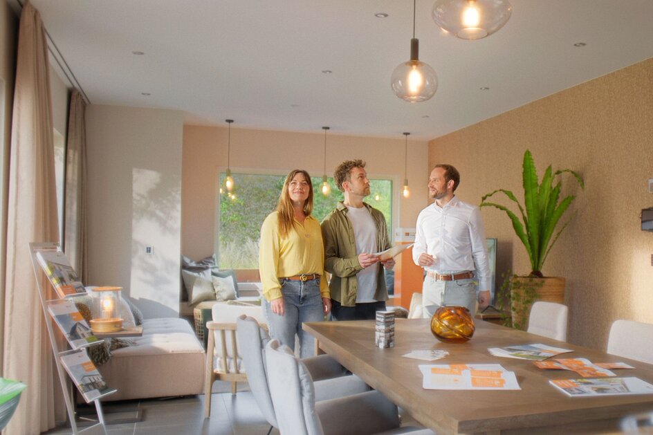 Heller Wohn- und Essbereich mit großem Holztisch, Polsterstühlen, Sofa und bodentiefen Fenstern; drei Personen stehen am Kopf des Tisches unter modernen Pendelleuchten, auf dem Tisch liegen Prospekte und Unterlagen, im Hintergrund sind Pflanzen und ein Sideboard zu sehen.