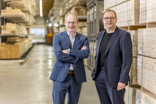 Zwei Männer in Anzügen stehen in einem Lager mit gestapelten Holzkisten und Paletten im Hintergrund - Geschäftsführer von FingerHaus Dr. Mathias Schäfer und Klaus Cronau