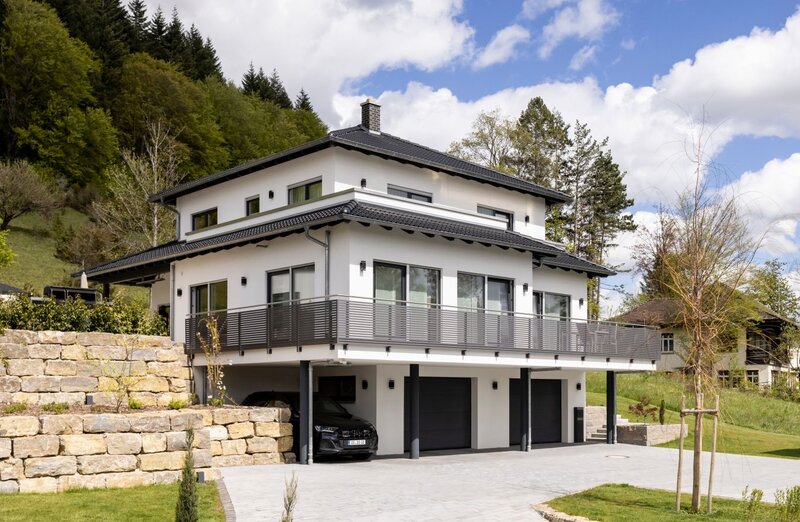 Frei geplantes Haus von FingerHaus mit mehrgeschossiger Architektur bei Tageslicht, helle Putzfassade, umlaufender Balkon mit Metallgeländer, integrierte Garage im Untergeschoss, gepflasterte Zufahrt, Stützmauern aus Naturstein und Hanglage mit Grünflächen.