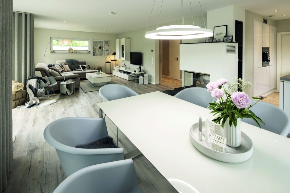 Offener Ess- und Wohnbereich mit großem Esstisch, grauen Stühlen und moderner Couch. Im Hintergrund Kamin und Fensterfront mit Vorhängen.