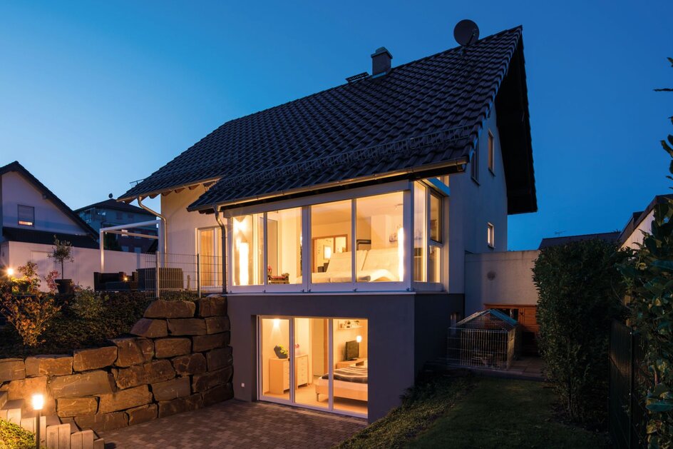 Fertighaus FingerHaus MEDLEY bei Abenddämmerung mit beleuchtetem Wohnbereich und ausgebautem Kellergeschoss mit großen Fenstern und Terrasse, eingebettet in Hanglage mit Natursteinmauer.