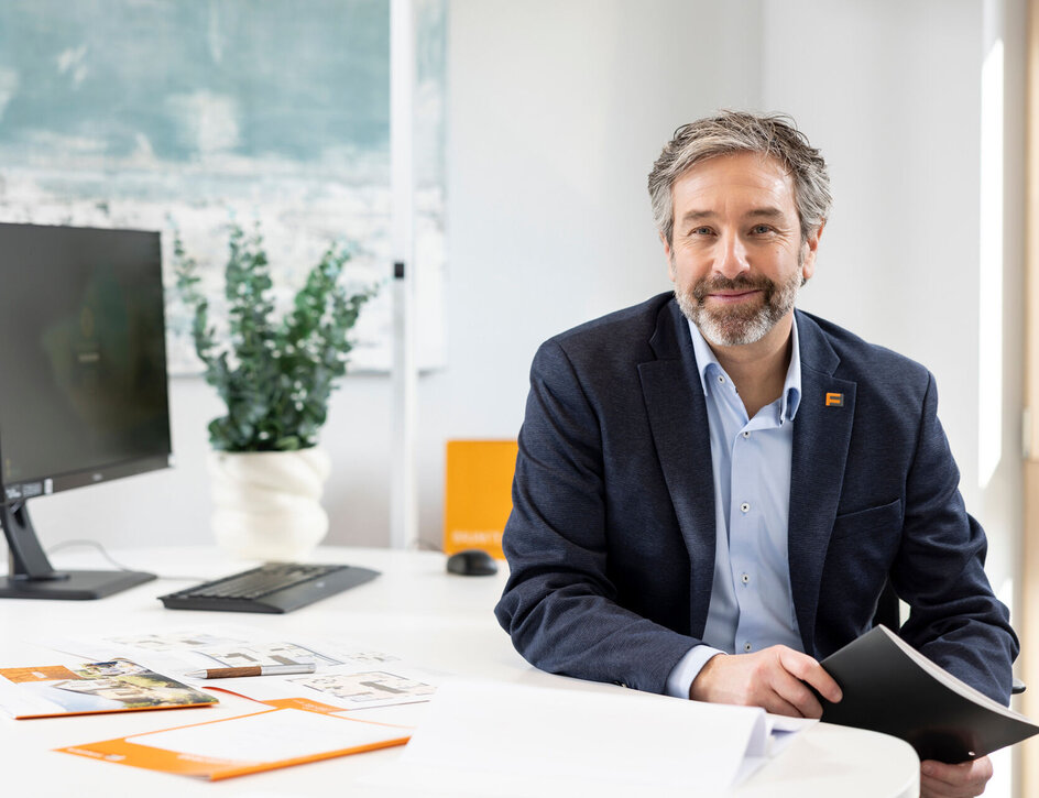 FingerHaus-Fachberater sitzt an seinem Schreibtisch und blickt freundlich in die Kamera, Unterlagen zur Hausplanung liegen vor ihm.