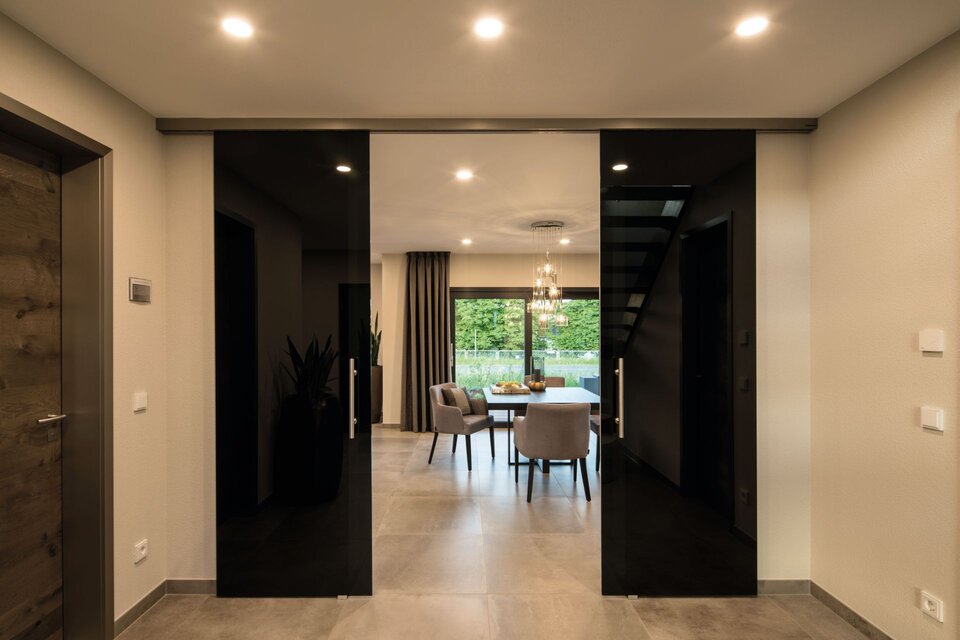 Blick durch einen Flur mit hellen Wänden und Boden auf einen Essbereich mit Tisch, Stühlen und großer Fensterfront, links eine Holztür, rechts eine Treppe, mittig zwei große schwarze Schiebetüren.