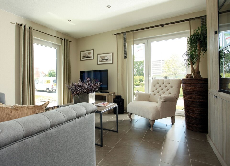 Helles Wohnzimmer im FingerHaus-Musterhaus MEDLEY 3.0 in Erfurt mit großer Fensterfront, gemütlichem Sofa, Sessel und Blick in den Garten.