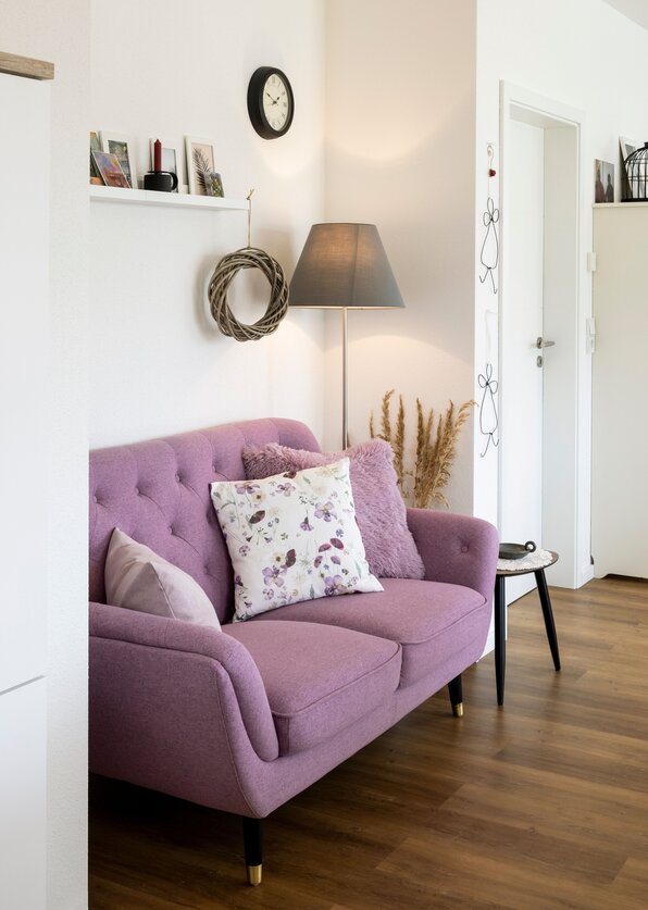 Wohnbereich mit rosafarbenem Sofa, Stehlampe, Wanduhr und dekorativen Elementen auf hellem Hintergrund