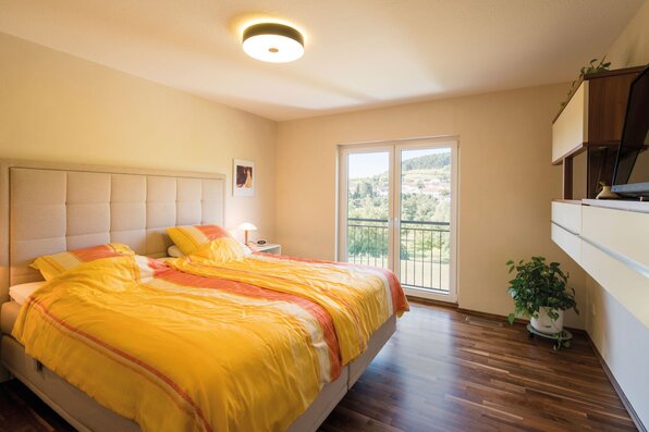 Helles Schlafzimmer in einem frei geplanten FingerHaus mit Doppelbett, gelber Bettwäsche, großen Fenstern mit Balkonzugang und Holzfußboden.