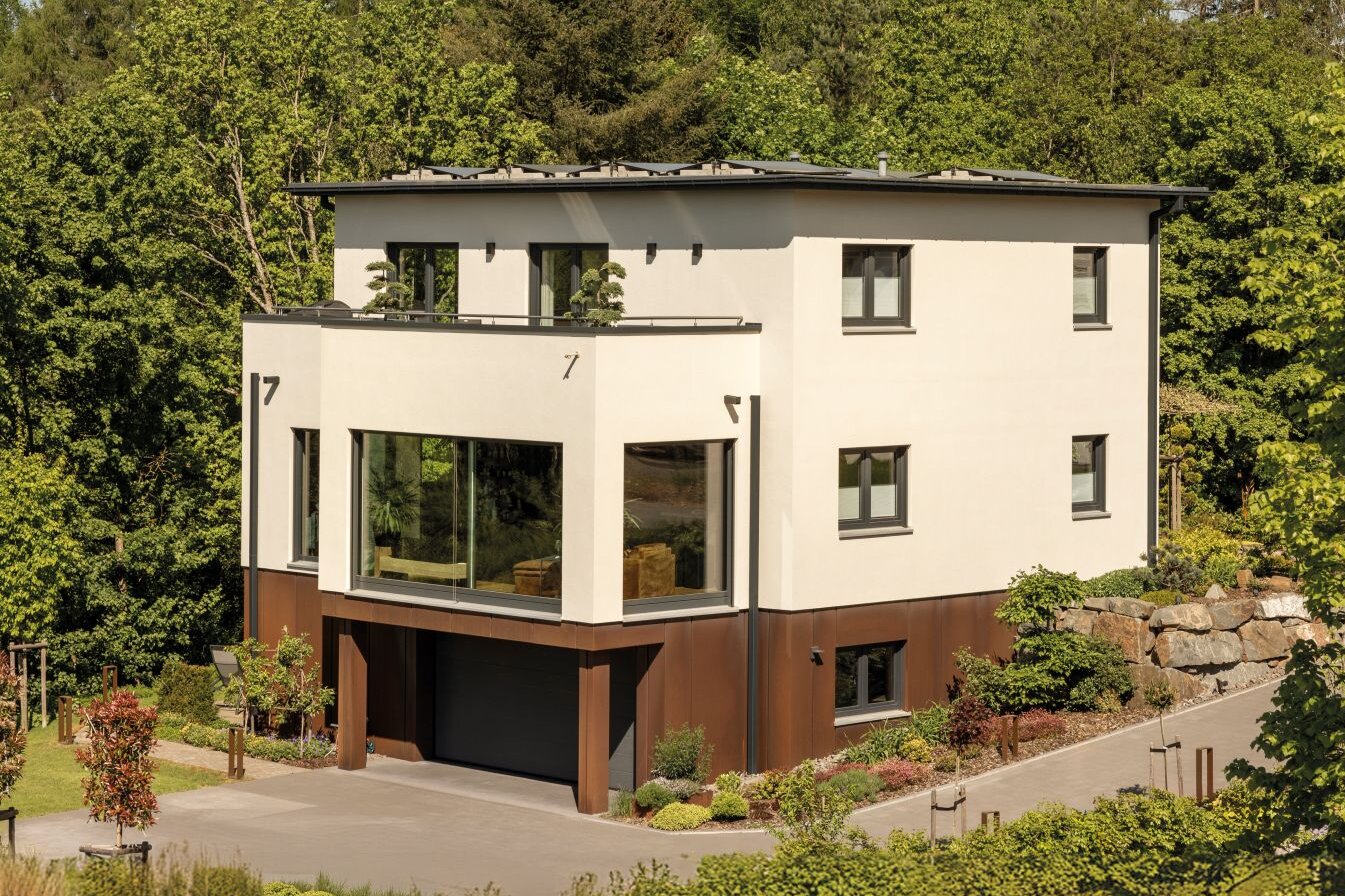 Mehrgeschossiges Wohnhaus mit heller Putzfassade, Flachdach, großen bodentiefen Fenstern, Balkon mit Geländer, integrierter Garage und hangseitigem Grundstück, umgeben von Bäumen.