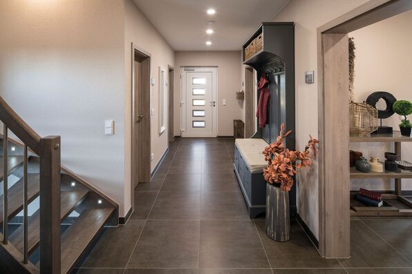 Geräumiger Flur mit dunklem Fliesenboden, Garderobe aus Holz mit Sitzbank und Ablagefächern im Musterhaus UNO in Leipzig. Im Hintergrund moderne Haustür mit Glaseinsätzen, links Holztreppe mit Beleuchtung und an der Wand dekorative Elemente.