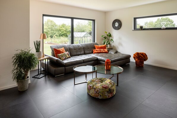 Helles Wohnzimmer mit grauer Eckcouch, farbigen Kissen, runden Couchtischen und großen Fenstern mit Blick in den Garten.