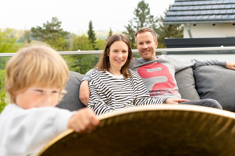 Ein Paar sitzt entspannt auf dem Sofa auf der Terrasse ihres modernen FingerHaus-Hauses, während im Vordergrund eines ihrer Kinder spielt.