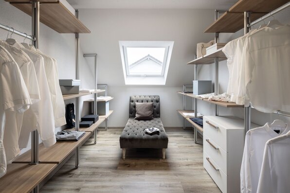 Ankleidezimmer mit offener Regallösung, Kleiderstangen und stilvoller Liege unter einem Dachfenster im Musterhaus VIO Köln.