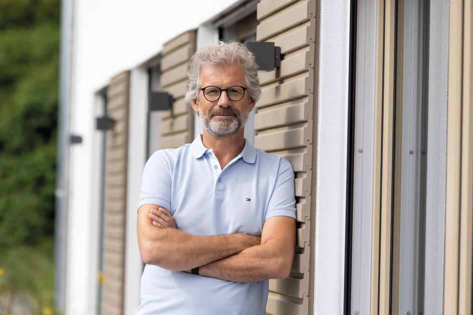 Mann mit verschränkten Armen steht vor der Fassade eines Finger-Hauses.