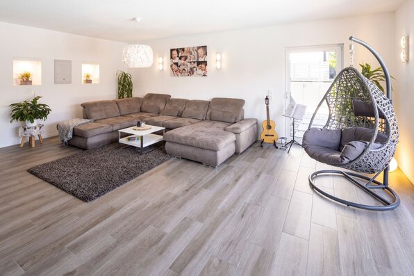 Helles Wohnzimmer in einem frei geplanten Finger-Haus mit großer Sofalandschaft, bodentiefem Fenster und modernem Bodenbelag.