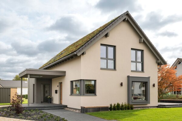 Frontansicht des Musterhauses JUNO 300 in Mülheim-Kärlich mit begrüntem Satteldach, überdachtem Eingang und großem Fenster im Erdgeschoss.
