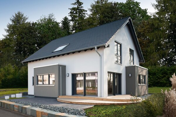 Modernes Musterhaus mit Satteldach und zweifarbiger Putzfassade in Weiß und Grau. Vor dem Haus befindet sich eine Holzterrasse, eingerahmt von Zierkies und einer niedrigen Natursteinmauer.