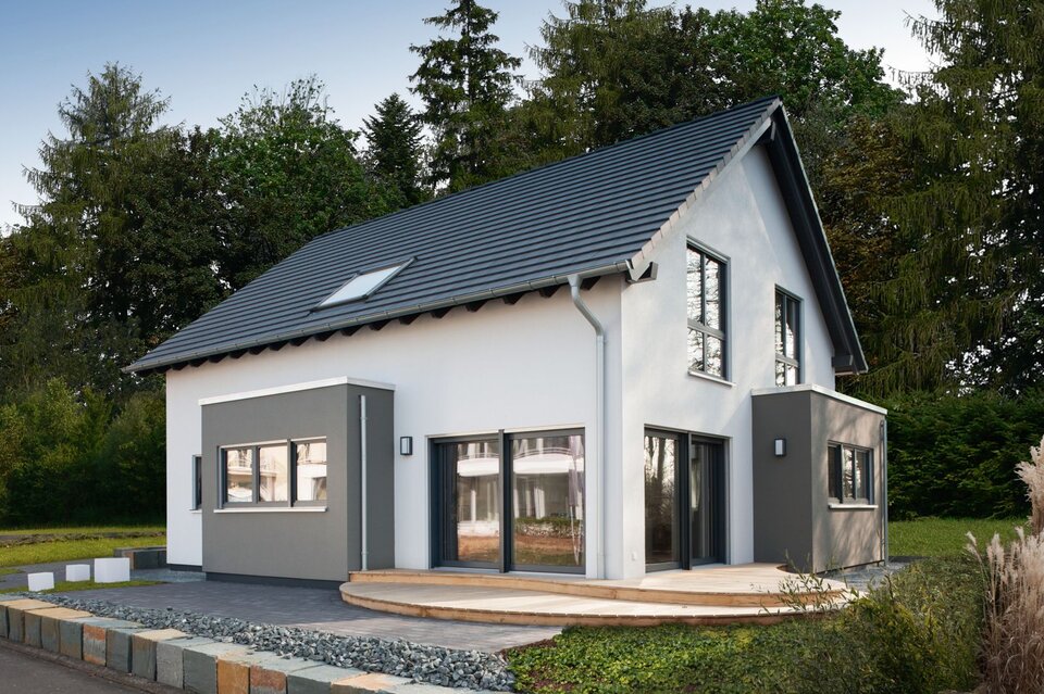 Modernes Musterhaus mit Satteldach und zweifarbiger Putzfassade in Weiß und Grau. Vor dem Haus befindet sich eine Holzterrasse, eingerahmt von Zierkies und einer niedrigen Natursteinmauer.