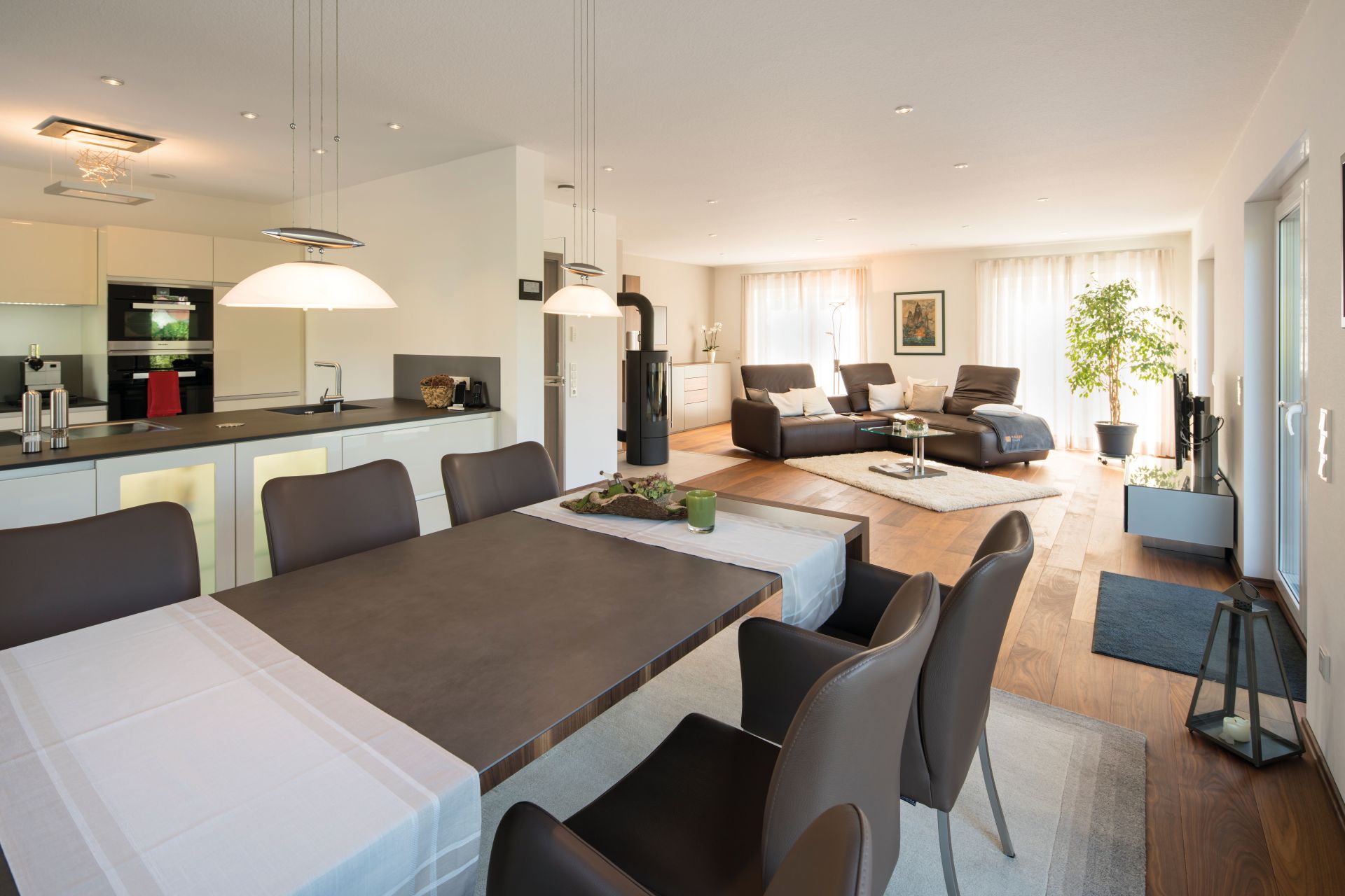 Offener Wohn- und Essbereich in einem frei geplanten FingerHaus mit moderner Küche, großem Esstisch, braunen Stühlen, gemütlicher Sofalandschaft und großen Fenstern mit viel Tageslicht.